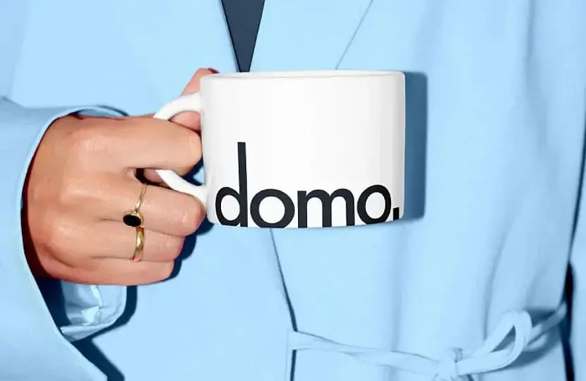 Close-up of a hand with gold rings holding a white Domo-branded mug against a pale blue jacket.
