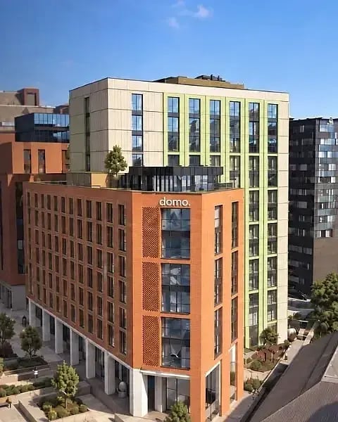 Exterior of the Domo Leeds student accommodation building, a modern red brick and cream block with green window detailing and a rooftop terrace, set among city buildings.