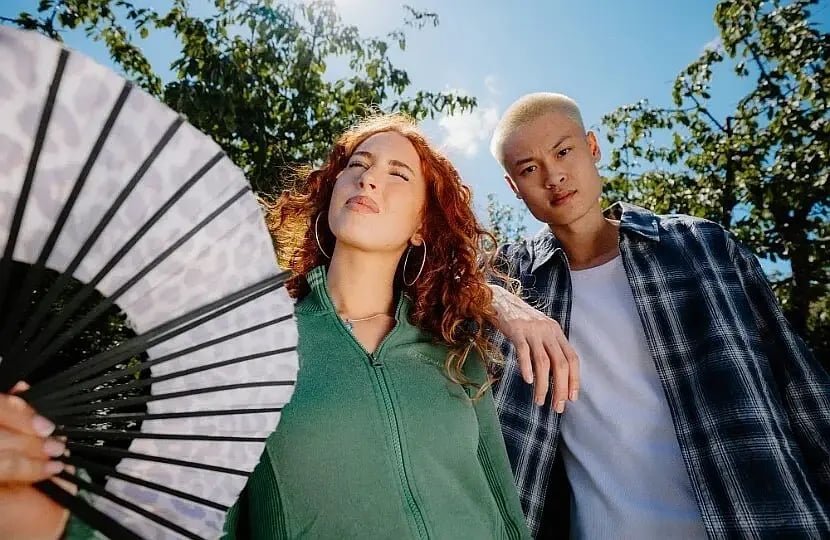 Two students posing outdoors under trees on a sunny day, one holding a large patterned hand fan, the other resting an arm on her shoulder.