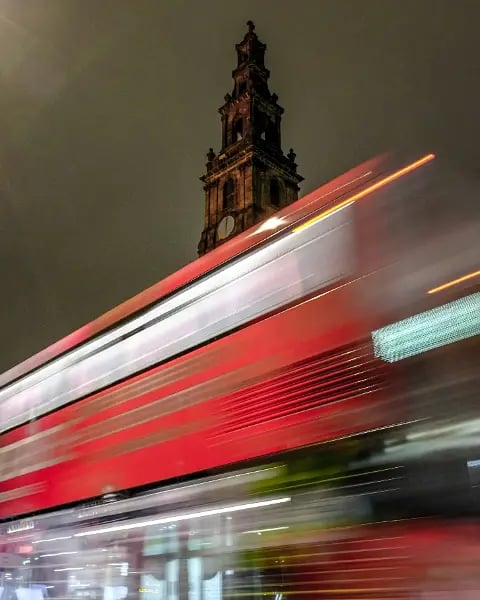 Red double-decker bus speeding past in a motion blur at night, beneath a floodlit church tower in Leeds city centre.