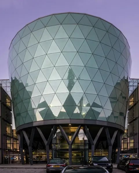 The Rose Bowl building at Leeds Beckett University, a curved glass bowl-shaped structure on angled supports, lit up at dusk.