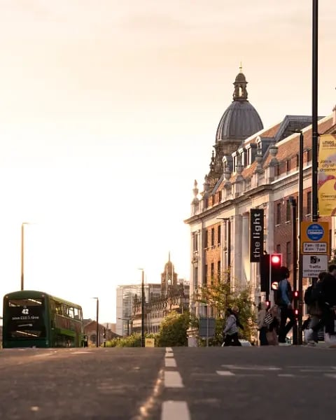 Leeds city centre street at golden hour, with a green double-decker bus, pedestrians crossing and the domed building of The Light shopping centre.