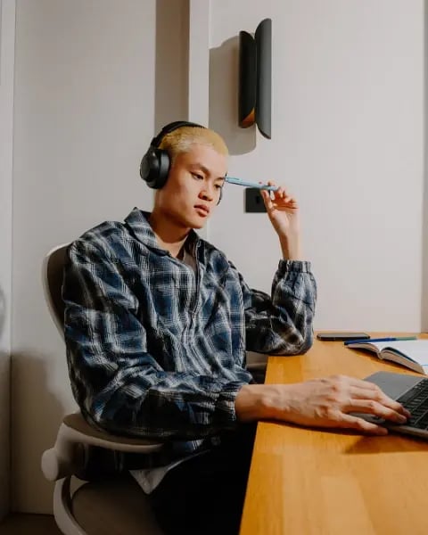 Student wearing headphones and a checked shirt studying at a wooden desk with a laptop and open notebook.