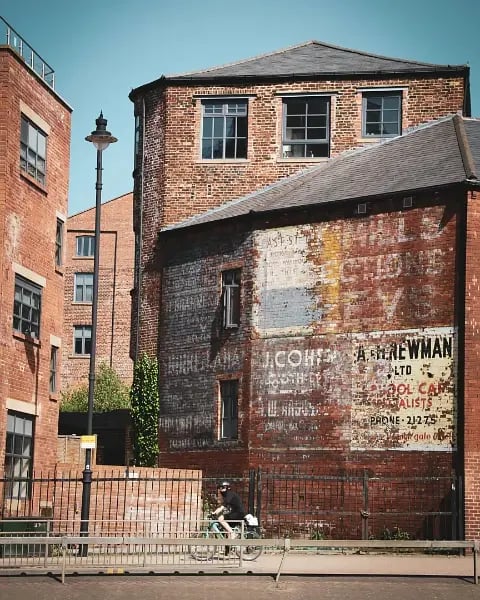 Cyclist riding past old red brick buildings in Leeds city centre, with faded painted advertisements on the side wall.